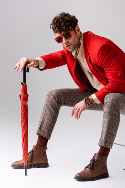 Модный мужчина в красном пиджаке наклоняется вперед и позирует с зонтиком, сидя на стуле на сером - Фото, изображение