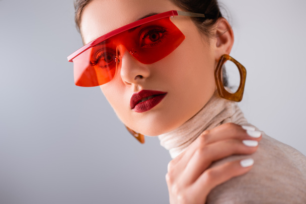 Attractive girl in red sunglasses touching shoulder and looking at camera isolated on grey  - Zdjęcie, obraz