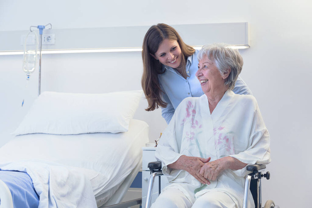 Gondozó vagy fiatal lány, kerekesszékben ülő idős nőt tolva, aki lehajol, hogy belsejében mosolyogjon egy kórházi szobában, kórteremben vagy öregek otthonában. - Fotó, kép
