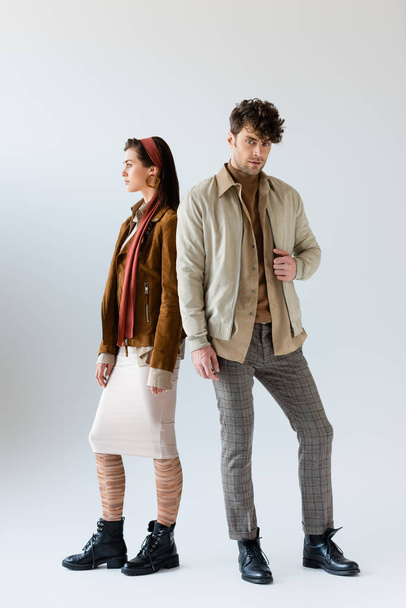 Volledige weergave van stijlvolle man in de herfst outfit kijken naar camera, staan in de buurt van aantrekkelijke vrouw weg te kijken op grijs - Foto, afbeelding