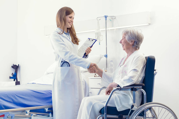Χαμογελώντας φιλική γυναίκα γιατρός σφίγγει το χέρι με μια ηλικιωμένη ασθενή κάθεται σε μια αναπηρική καρέκλα στο δωμάτιό της στο νοσοκομείο κατά τη διάρκεια θαλάμων - Φωτογραφία, εικόνα