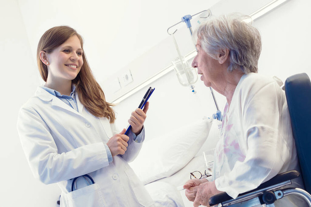 Νεαρή, φιλική, χαμογελαστή γυναίκα, ιατρός, σύμβουλος σε ηλικιωμένη γυναίκα ασθενή που κάθεται σε αναπηρική καρέκλα κρατώντας σημειώσεις κατά τη διάρκεια της εξέτασης σε εσωτερικούς χώρους στην πτέρυγα του νοσοκομείου - Φωτογραφία, εικόνα