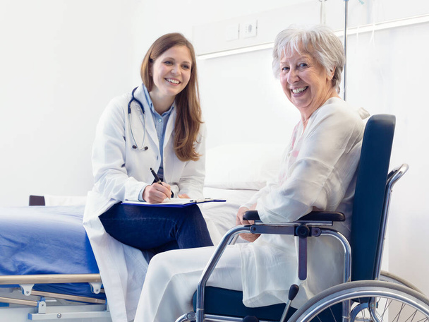 Verpleegster of arts die patiëntenaantekeningen schrijft op een afdeling voor een glimlachende oudere vrouw die in een rolstoel zit in een medisch en gezondheidsconcept - Foto, afbeelding