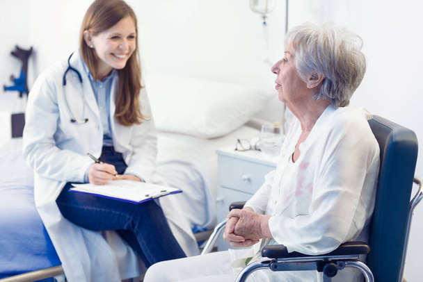 Infermiera o medico che scrive note di paziente in un reparto per una donna anziana sorridente seduta su una sedia a rotelle in un concetto medico e sanitario - Foto, immagini