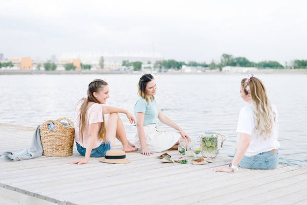 three girls sitting on a wooden pier in front of the water. rest, picnic,lemonade - Foto, afbeelding