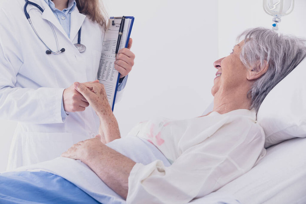 Χαμογελώντας γυναίκα γιατρός ή νοσοκόμα παρηγορεί μια ηλικιωμένη γυναίκα ασθενή σε ένα κρεβάτι που απλώνεται για να κρατήσει το χέρι της κατά τη διάρκεια γύρων σε ένα νοσοκομείο - Φωτογραφία, εικόνα