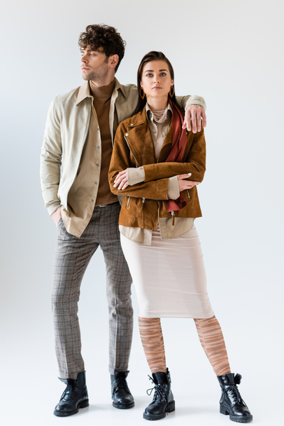 Полный обзор стильного мужчины с рукой в кармане, глядя в сторону, обнимая привлекательную женщину, стоящую рядом на сером - Фото, изображение