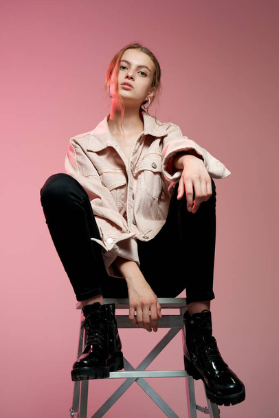 A girl in a pink jeans in black jeans sits on a white wooden bar stool and poses on a pink background. - Foto, imagen