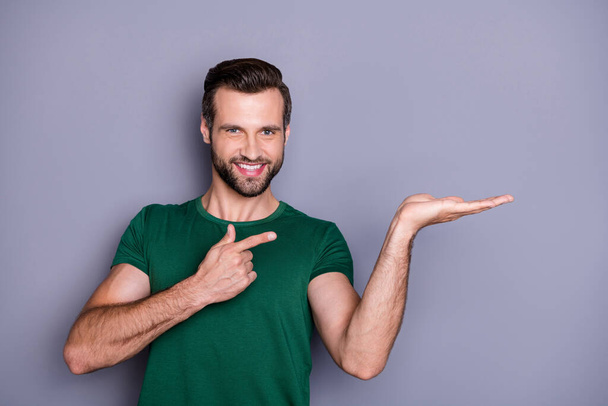 Photo of attractive handsome guy good mood direct finger side empty space open arm holding cool offer shopping product wear casual green t-shirt isolated grey color background - Foto, Imagem