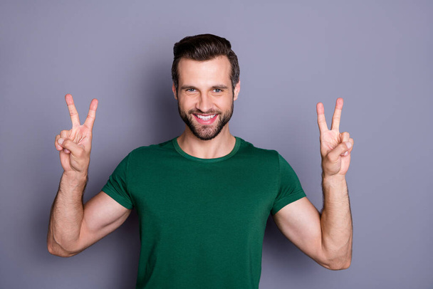 Photo of attractive handsome bearded guy raise hands showing v-sign symbols fingers saying hi to friends wear casual green t-shirt isolated grey color background - Photo, image