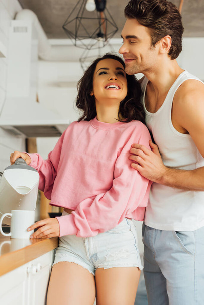 Young man hugging smiling woman pouring water in cup in kitchen  - Foto, Bild