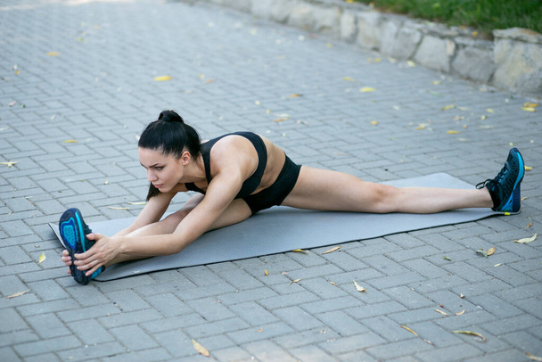 Slim young sportswoman with the athletic body does stretching exercises on the mattress for gymnastics outside in the park. - Photo, Image