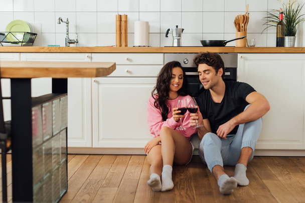 foco selectivo de sonriente pareja tintineo con vino en el suelo en la cocina
  - Foto, Imagen