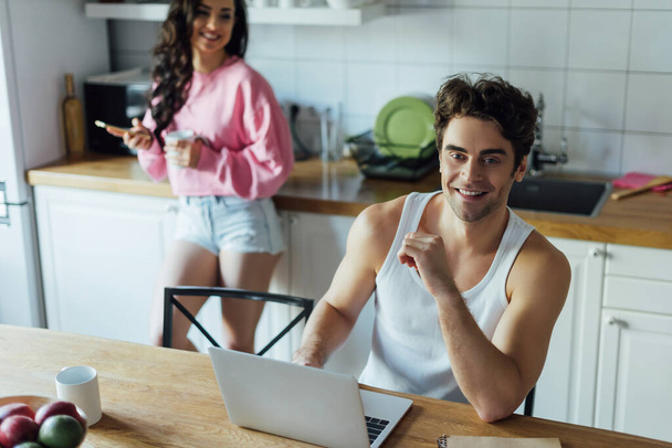 Enfoque selectivo del hombre sonriente utilizando el ordenador portátil cerca de la novia con taza y teléfono inteligente en la cocina
  - Foto, imagen