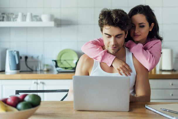 Selective focus of woman embracing boyfriend using laptop in kitchen  - Фото, изображение