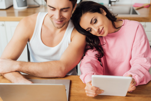 Selective focus of smiling man using laptop near beautiful girlfriend with digital tablet in kitchen  - Φωτογραφία, εικόνα
