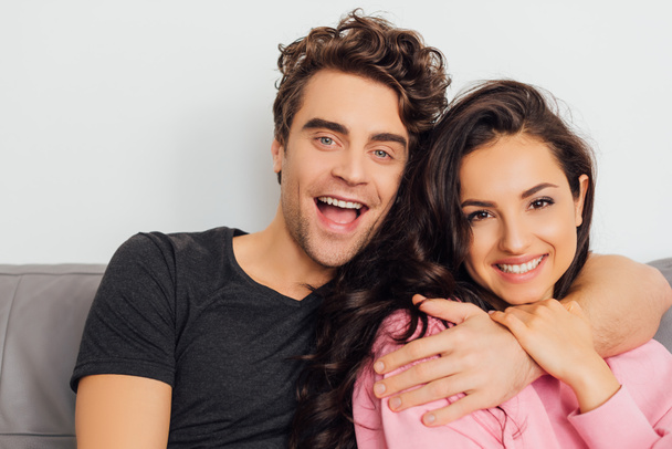 Young man embracing smiling girlfriend while looking at camera on couch on grey background - Foto, afbeelding