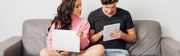 Panoramaorientierung des jungen Paares mit digitalem Tablet und Laptop auf der Couch auf grauem Hintergrund - Foto, Bild
