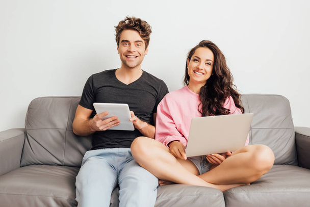 Νεαρό ζευγάρι χαμογελά στην κάμερα, ενώ κρατώντας φορητό υπολογιστή και ψηφιακό tablet στον καναπέ σε γκρι φόντο  - Φωτογραφία, εικόνα