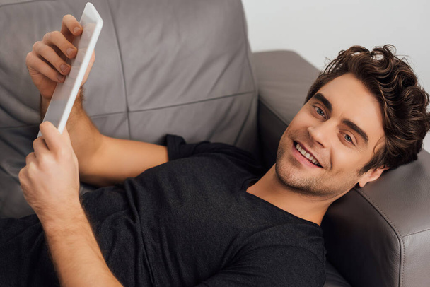 Smiling man looking at camera while holding digital tablet on couch isolated on grey - Foto, immagini