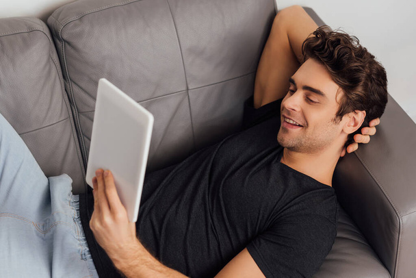 Foco seletivo de homem bonito sorrindo enquanto segura tablet digital no sofá em fundo cinza
  - Foto, Imagem