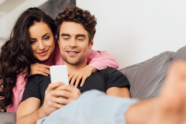 Selective focus of smiling man using smartphone near beautiful girlfriend at home  - Fotó, kép