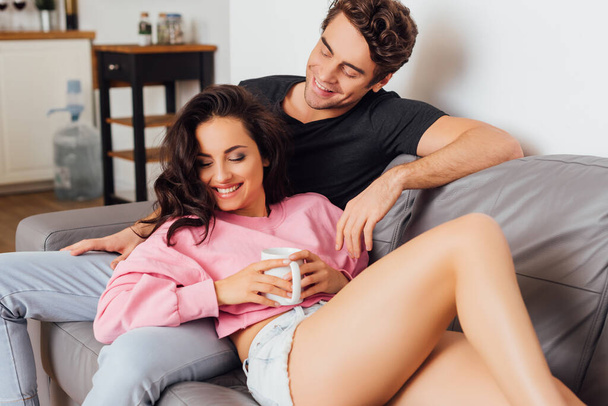 Smiling woman holding cup of coffee on couch near boyfriend in living room - Foto, Imagem