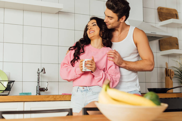 Selective focus of man embracing beautiful smiling girlfriend with cup of coffee in kitchen  - Valokuva, kuva