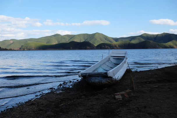 un hilo de barco blanco en la playa de suelo negro. Olas azules del lago alrededor. Fondo de montañas verdes. cielo azul soleado. En Lugu lago Sichuan China.  - Foto, Imagen
