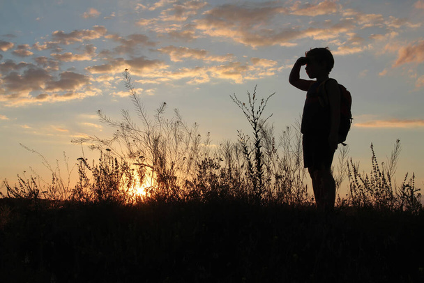 Bébé garçon avec un sac à dos regarde le coucher de soleil avec sa paume dans les yeux - Photo, image