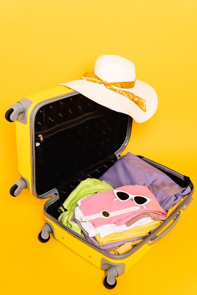 nyitott utazási táska nyári ruhák és kiegészítők sárga háttér - Fotó, kép