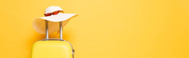 žlutá cestovní taška se slunečním kloboukem a slunečními brýlemi izolovaná na žlutém, panoramatickém snímku - Fotografie, Obrázek
