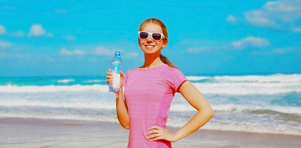Fitness happy woman drinking water from bottle on the beach near the sea - 写真・画像
