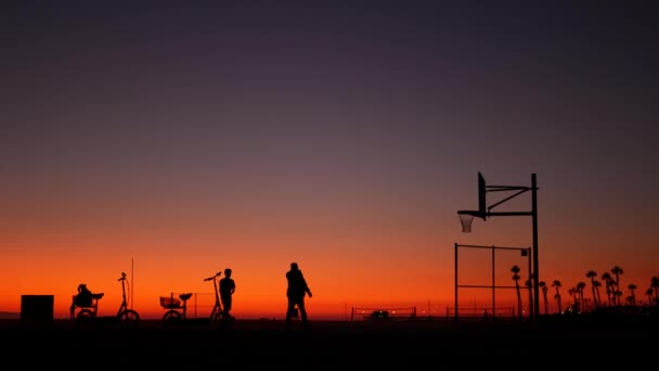カリフォルニアの夏の夕暮れのビーチの美しさ、ピンクの夕日。認識できないシルエット、人々はバスケットボールコートでボールでゲームをプレイします。ロサンゼルスの近くのニューポート海洋リゾートCA USA 。紫色の空のグラデーション - 映像、動画