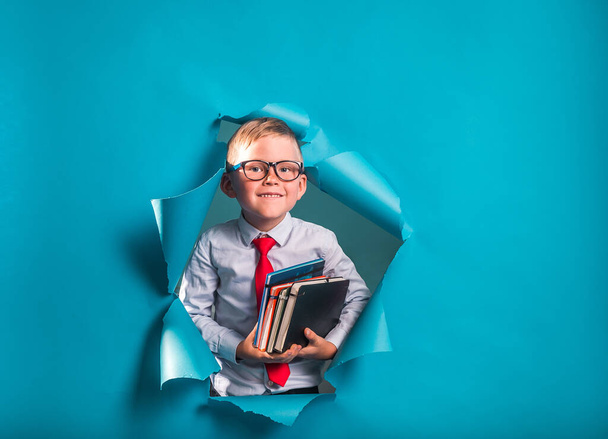 Vissza a suliba. Szorgalmas kisfiú hátizsákkal és könyvekkel. Happy kölyök áttöri a papírfalat. Vicces tanuló szemüvegben felfelé mutat. - Fotó, kép