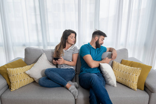 Young boyfriend making fun and jealous his girlfriend pretending that he is texting with other girl on his cell or mobile phone while they sitting on the sofa in their apartment - Фото, изображение