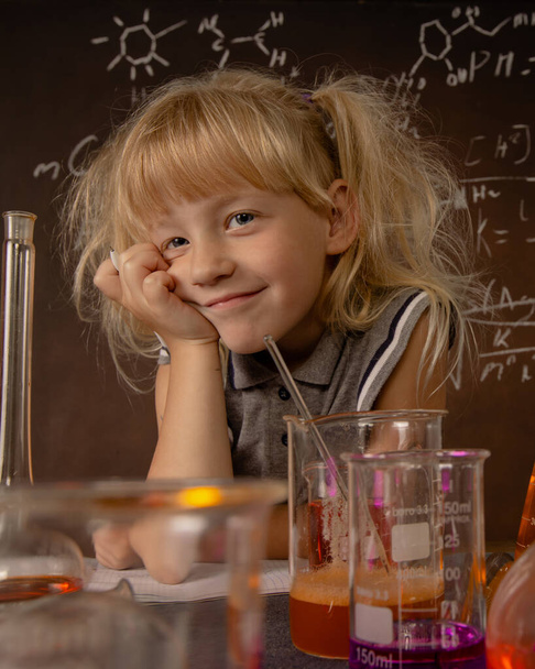 Curious little girl with test tubes and colorful substances with hand to her face makes tests at school laboratory. Small kid conducts science experiment on biology lesson. Biology education concept. - Photo, Image