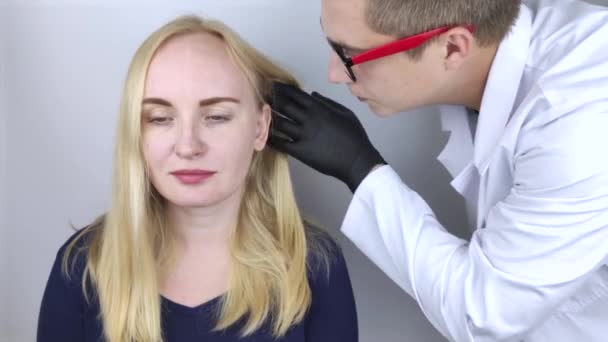 Lääkäri trikologi tekee tutkimuksen blondin hiuksista. Hiustenlähtö hoito. Hiustenlähtö, hiustenlähtö, kutina, pään polttelu, seborrea tai hauraat päät - Materiaali, video