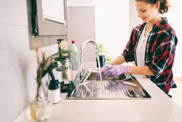 Young charming happy smiling girl woman washing dishes in gloves in the kitchen. House Hygiene and Cleaning Service Concept. - Foto, afbeelding