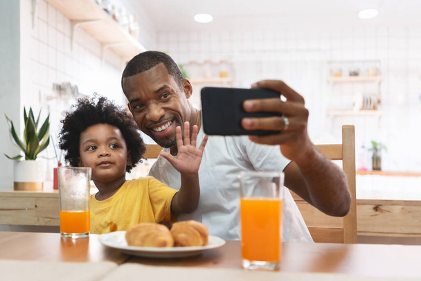 Happy African-American man and kid boy using mobile phone for taking selfie together at home. Cheerful African Father and son doing video chat and waving hand during talking to family with smartphone - Foto, Bild