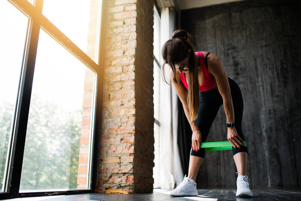 Untere Ansicht eines Mädchens korrigiert ein Gummi-Sportband, das an den Waden getragen wird, um die inneren Muskeln der Oberschenkel in einem Loft zu trainieren - Foto, Bild