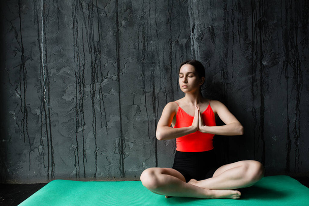 Photo of a girl fitness trainer meditating on a sports mat in a loft with her arms folded in front of her in a lotus position - Foto, Bild