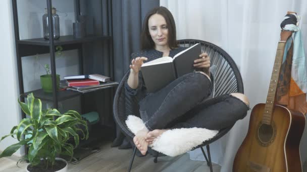 Young woman sitting on cozy chair, relaxing and reading book. Relaxation at home. Girl at home sitting on chair in front of window and reading book. Carefree female reading book sitting on chair - Filmagem, Vídeo
