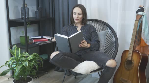 Carefree brunette woman in earphones sitting on cozy chair, relaxing, listening to music and reading book. Relax time at home. Smiling dreaming girl relaxing in chair at modern apartment in evening - Πλάνα, βίντεο