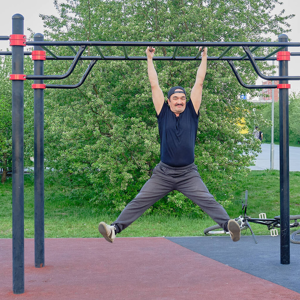Happy senior asian man is training on the playground in the park on the background of a flowering plant. Hanging on the bar. Life style - Photo, Image