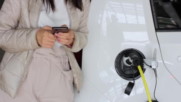 Mujer con un teléfono inteligente cerca de recargar el coche eléctrico y comprobar el estado de la batería. Carga de vehículos en la estación de carga pública al aire libre. Concepto de coche compartido
 - Imágenes, Vídeo