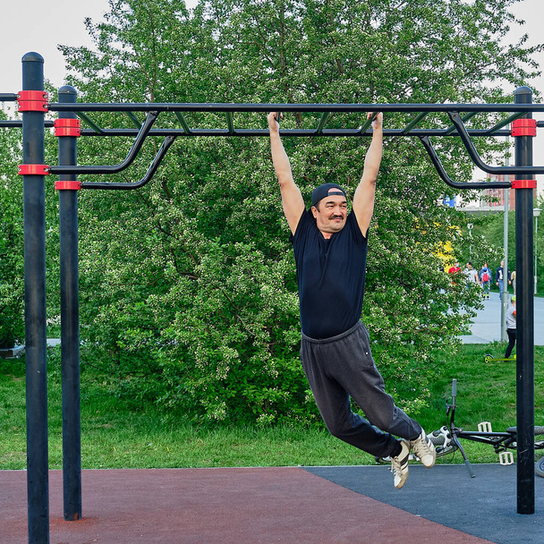 Happy senior asian man is training on the playground in the park on the background of a flowering plant. Hanging on the bar. Life style - Foto, Imagem