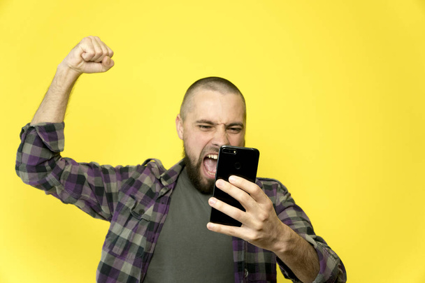 Ein junger Mann mit Bart und Glatze hält auf gelbem Hintergrund ein Handy in der Hand. Ein Mann hält überglücklich die Hand in der Faust über dem Kopf - Foto, Bild