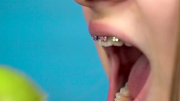 Dişlerinde diş teli olan genç bir kız mavi arka planda yeşil bir elma yiyor. Renkli diş telleri olan bir kız elmanın yakın çekimini ısırıp koparır.. - Video, Çekim
