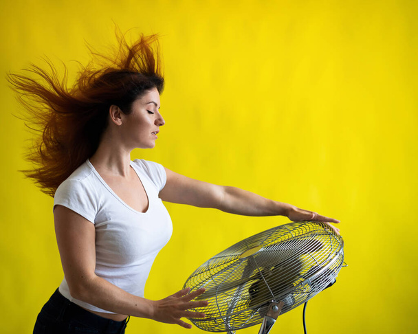 A beautiful red-haired woman is cooled off standing over a large electric fan on a yellow background. Girl with hair developing in the wind. Device for cooling the air. - Foto, Bild